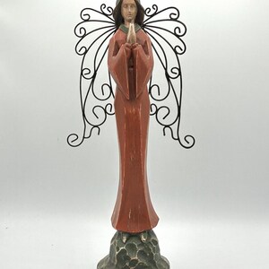 May include: A standing angel figurine with a terracotta-colored robe, dark hair, and hands pressed together in prayer. The figure features black wire wings with a scrollwork design and a matching wire halo, all mounted on a textured dark green base.