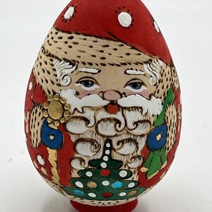 May include: Hand-painted wooden egg ornament depicting Santa Claus. The egg features a red hat with white polka dots, a white beard, and a green Christmas tree design. The ornament is approximately 4 inches tall.