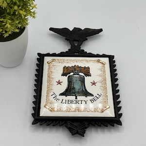 Könnte beinhalten: Schwarze Liberty Bell Wanddekoration aus Metall mit einem Adler. Die quadratische Fliese zeigt die Liberty Bell, zwei rote Sterne und die Worte "The Liberty Bell" in einem dekorativen Rahmen.
