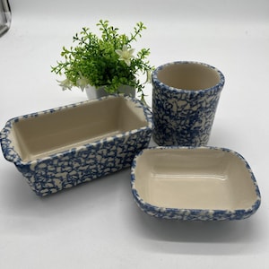 Puede incluir: Un conjunto de utensilios de cocina de cerámica con un patrón azul y blanco. El juego incluye una fuente rectangular para pan, un plato cuadrado pequeño y una taza cilíndrica. El interior de cada pieza es de color crema.