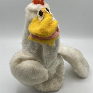 以下が含まれることがあります： 白い体、黄色のくちばし、オレンジ色の鶏冠を持つ、ぬいぐるみニワトリの人形。人形は黒い目とくちばしの周りにピンクのアクセントがあります。ニワトリの翼は広げられており、柔らかくふわふわした素材でできているようです。