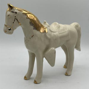 Puede incluir: Figura de caballo de cerámica en posición de pie. El caballo es principalmente blanco roto con detalles dorados en la crin, las pezuñas y la brida. También se ve una silla de montar. La figura tiene un acabado brillante.