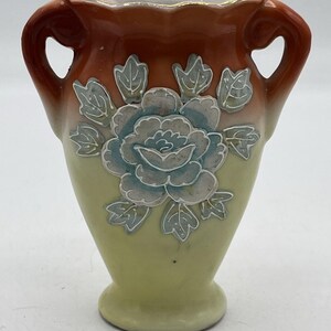 May include: A small, decorative vase with two handles. The vase has a light green base that fades to a brown top. A large, light blue floral design is centered on the vase, with white accents. The rim is trimmed with gold.