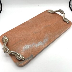 May include: A rectangular tray made of pink stone with a smooth, polished surface. The tray features two silver-toned metal handles, each designed with a textured, twisted rope pattern attached to opposite ends of the stone base.