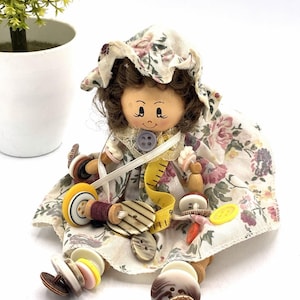 May include: A doll with brown hair wearing a floral print bonnet and dress, adorned with various colorful buttons and a yellow measuring tape. The doll features a painted face and is constructed with wooden limbs, sitting amidst a collection of buttons.