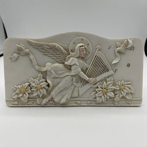 May include: A rectangular, off-white angel figurine playing a harp. The angel has large wings and a halo. Poinsettias with yellow centers and doves decorate the base. Musical notes are also present.