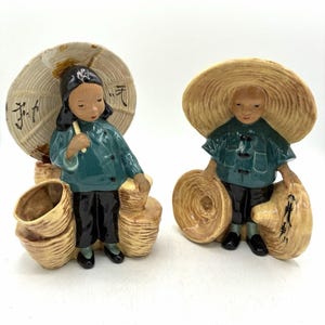 May include: Two ceramic figurines depicting individuals wearing teal shirts, black trousers, and wide-brimmed straw hats. One figure holds a large umbrella with black calligraphy, while both figures are surrounded by woven baskets with textured details.