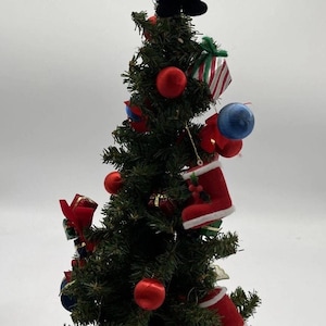 May include: A small Christmas tree adorned with red baubles, miniature gift boxes, and red and white Santa boots. The tree is topped with a teddy bear dressed as a drummer. The tree is set on a small, natural-looking base.