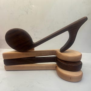 Puede incluir: Utensilios de cocina de madera con forma de nota musical. El conjunto incluye una cuchara de madera marrón oscuro y varios elementos de madera marrón claro y oscuro con forma de nota. Los utensilios están apilados, mostrando la veta natural de la madera.