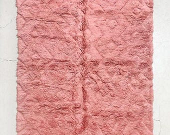 May include: A rectangular, plush, pink rug with a textured surface and diamond pattern. The rug has a white fringe along the bottom edge. The rug is displayed on a light gray concrete surface.