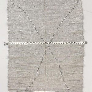 Peut inclure: Un tapis rectangulaire gris avec une texture tissée. Le tapis présente des bordures blanches avec des motifs à points noirs et des lignes diagonales cousues en noir et blanc. De petits pompons sont visibles aux coins.