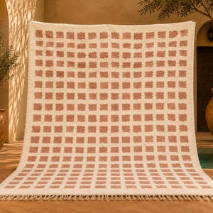 Peut inclure: Un tapis rectangulaire présentant un motif de grille répétitif composé de carrés couleur terre cuite sur un fond crème. Le tapis possède une surface à poils touffetés texturés et est fini avec une frange sur le bord inférieur.