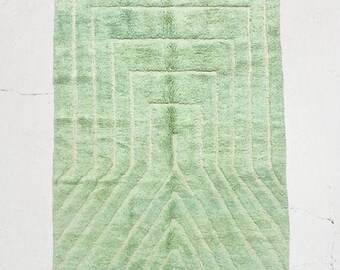 May include: A rectangular, pale green rug with a geometric pattern. The rug has a series of nested rectangles and angled lines in a lighter shade of green, with cream-colored fringe along the edges. The rug is designed for home decor.