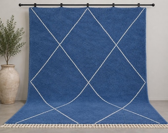 Puede incluir: Una alfombra rectangular azul con un patrón de rombos blancos en toda su superficie. La alfombra tiene un pelo grueso y texturizado, rematada con flecos cortos de color crema en el borde inferior, colgada de una barra de metal negro.