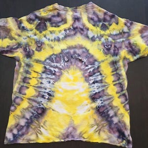 Pode incluir: Uma camiseta tie-dye com um padr&atilde;o vibrante. A camisa apresenta um design amarelo central que irradia para fora, complementado por detalhes roxos e brancos. O efeito geral &eacute; um design abstrato colorido.