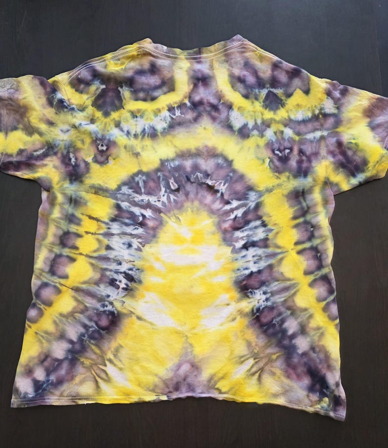 Pode incluir: Uma camiseta tie-dye com um padr&atilde;o vibrante. A camisa apresenta um design amarelo central que irradia para fora, complementado por detalhes roxos e brancos. O efeito geral &eacute; um design abstrato colorido.