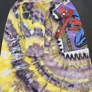 Pode incluir: Camiseta tie-dye de manga comprida com um gr&aacute;fico do Homem-Aranha. A camisa apresenta um padr&atilde;o tie-dye vibrante em amarelo, roxo e branco. O gr&aacute;fico do Homem-Aranha est&aacute; em um fundo roxo com uma paisagem urbana.
