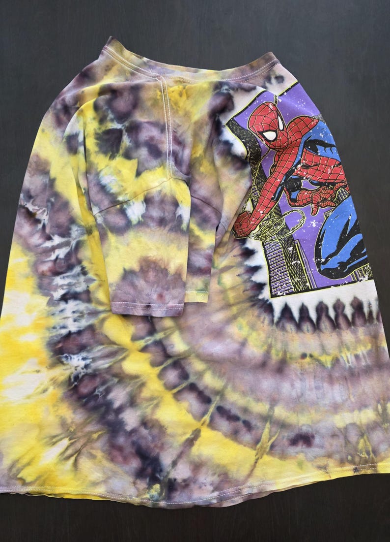 Pode incluir: Camiseta tie-dye de manga comprida com um gr&aacute;fico do Homem-Aranha. A camisa apresenta um padr&atilde;o tie-dye vibrante em amarelo, roxo e branco. O gr&aacute;fico do Homem-Aranha est&aacute; em um fundo roxo com uma paisagem urbana.