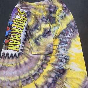 Pode incluir: Camiseta tie-dye de manga comprida com um padr&atilde;o espiral amarelo, roxo e branco. A manga esquerda apresenta um gr&aacute;fico do Homem-Aranha com o texto "SPIDER-MAN" em amarelo.