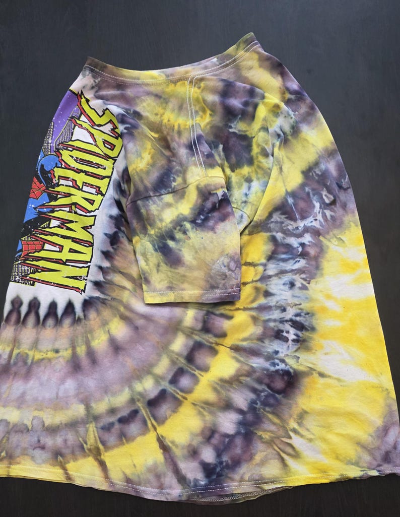 Pode incluir: Camiseta tie-dye de manga comprida com um padr&atilde;o espiral amarelo, roxo e branco. A manga esquerda apresenta um gr&aacute;fico do Homem-Aranha com o texto "SPIDER-MAN" em amarelo.