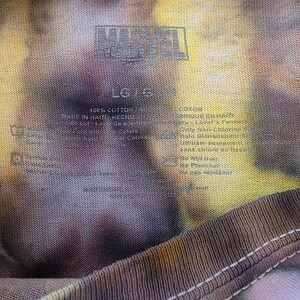 Pode incluir: Close-up de uma camiseta tie-dye com o logotipo da Marvel. A camisa &eacute; uma mistura de cores, incluindo amarelo, roxo e marrom. A etiqueta indica que &eacute; 100% algod&atilde;o e tamanho LG.