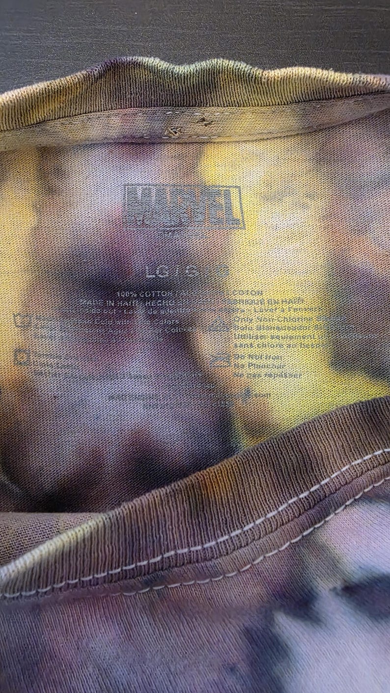 Pode incluir: Close-up de uma camiseta tie-dye com o logotipo da Marvel. A camisa &eacute; uma mistura de cores, incluindo amarelo, roxo e marrom. A etiqueta indica que &eacute; 100% algod&atilde;o e tamanho LG.