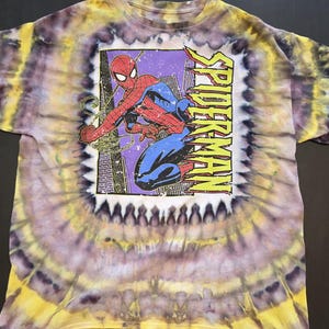 Pode incluir: Camiseta tie-dye com um gr&aacute;fico do Homem-Aranha. A camisa tem um padr&atilde;o espiral amarelo, roxo e branco. O gr&aacute;fico do Homem-Aranha est&aacute; em uma moldura retangular com a palavra "SPIDERMAN" em amarelo.