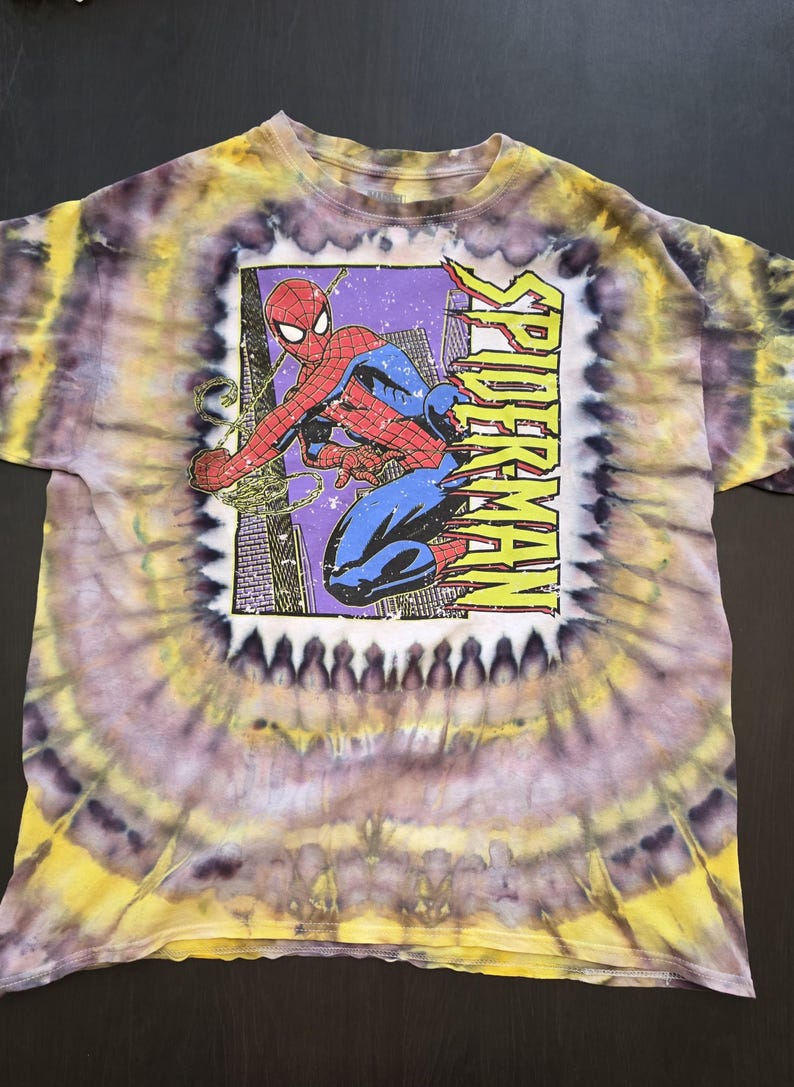 Pode incluir: Camiseta tie-dye com um gr&aacute;fico do Homem-Aranha. A camisa tem um padr&atilde;o espiral amarelo, roxo e branco. O gr&aacute;fico do Homem-Aranha est&aacute; em uma moldura retangular com a palavra "SPIDERMAN" em amarelo.