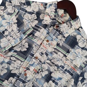 May include: A short-sleeved button-down shirt featuring a dark blue and white plaid pattern overlaid with a large white floral print. The shirt includes a classic collar, a front button placket with brown buttons, and thin green and red accent lines.