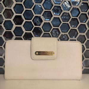 May include: A cream-colored wallet with a flap closure. The wallet features a gold-tone metal plate with the words "Juicy Couture" on the front. The background is a mosaic of blue and gray hexagonal tiles.