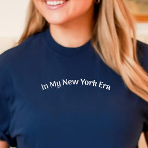 May include: A navy blue t-shirt with the text "In My New York Era" in white. The shirt has rolled-up sleeves and is made of a soft material. A cream-colored sweater is draped over the shoulders.