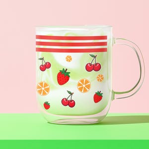 May include: A clear glass mug with a handle, filled with a green and white beverage. The mug is decorated with red stripes and fruit illustrations, including cherries, strawberries, and orange slices. The mug sits on a green surface against a pink backdrop.