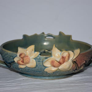 May include: A teal-colored ceramic bowl with two handles and floral accents. The bowl features two large, cream-colored flowers with peach centers. The rim has a decorative, scalloped edge. The bowl is approximately 10 inches in diameter.