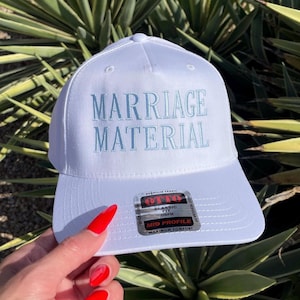 May include: A white baseball cap with the embroidered text "MARRIAGE MATERIAL" in light blue. The cap has a curved brim and a structured fit. The cap is a mid-profile style and is made of premium fabric.