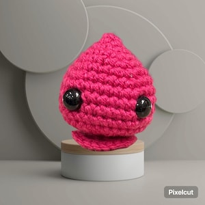 May include: A handmade, hot pink crocheted creature with large, black, glossy eyes. The amigurumi is displayed on a small, round wooden stand atop a white cylindrical base. The overall design is whimsical and cute.
