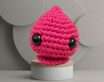 May include: A handmade, hot pink crocheted creature with large, black, glossy eyes. The amigurumi is displayed on a small, round wooden stand atop a white cylindrical base. The overall design is whimsical and cute.