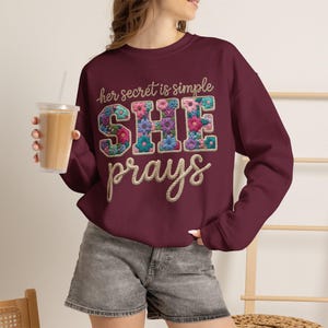 May include: A maroon long-sleeve crewneck sweatshirt featuring the text "her secret is simple SHE prays" in a rope-style font. The letters "SHE" are filled with a colourful floral pattern in shades of pink, blue, purple, and green.