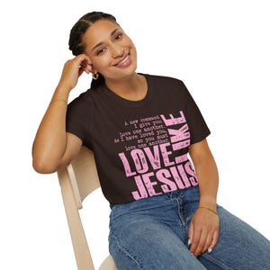 May include: A brown t-shirt with the text "LOVE JESUS" in distressed pink lettering. Above the text is a quote about love. The shirt is worn by a person wearing blue jeans.