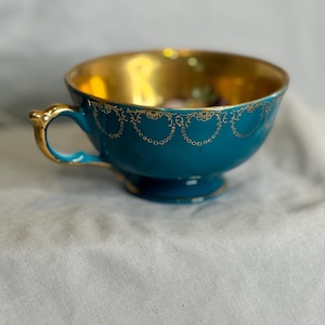 May include: Teal teacup with a gold interior and handle. The exterior features gold filigree detailing along the rim. The teacup sits on a small pedestal base. The handle is also gold.