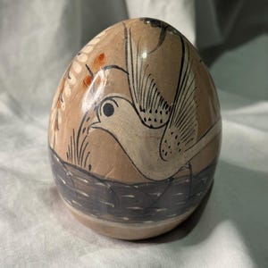 May include: A hand-painted, egg-shaped ceramic piece. The beige surface features a bird design with black outlines and details, including a wing and eye. The bird is above a dark gray band, with additional plant-like details.