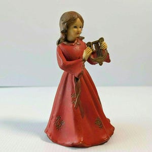 May include: A figurine of a person with brown hair wearing a long red dress with a tie waist and small floral patterns. The figure is holding a small, metallic-colored lyre instrument in both hands. The dress has long sleeves and a collar.