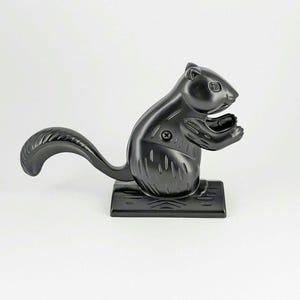 May include: A black squirrel-shaped nutcracker. The nutcracker is made of a shiny, dark material and features a long, curved tail. It has a square base and is designed to crack nuts.