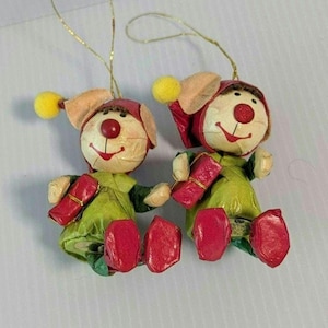 May include: Two hanging ornaments shaped like clowns, each wearing a red hat with a yellow pom-pom, a green outfit, and red shoes. Each figure has a round cream-colored face with a red nose and holds a small red rectangular gift box.