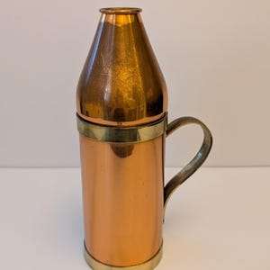 May include: A cylindrical copper vessel with a tapered top and a brass handle. The body features a polished copper finish, while the base, rim, and handle are constructed from brass. The vessel has a narrow opening at the top and a sturdy handle.