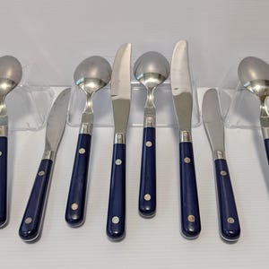 May include: A set of stainless steel silverware with navy blue handles. The set includes spoons and knives. The handles are a solid navy blue with a silver accent and a single silver rivet. The silverware is displayed on clear plastic stands.