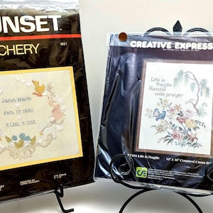 May include: Two counted cross stitch kits in plastic packaging. The left kit, Sunset Stitchery, features a baby keepsake design with rabbits and ducks. The right kit, Creative Expressions, displays a floral design with the text Life is fragile Handle with prayer.