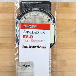 May include: An AirClassics E6-B Flight Computer, with instructions, is displayed in a clear plastic sleeve. The computer has a circular design with degree markings and a rotating dial. The text "For Ground Speed and True Heading" is visible.