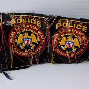 May include: Four black police patches with gold, red, and blue embroidery. Each patch features the word "POLICE" at the top, a circular emblem with an eagle, and text that reads "WHERE S.C. HISTORY OF GEORGETOWN BEGAN FOUNDED IN 1734."