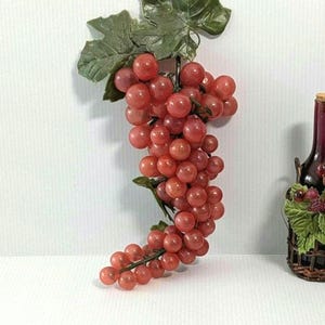 May include: A decorative cluster of artificial red grapes featuring numerous round, glossy berries attached to a central stem with several textured green leaves at the top. The grapes are arranged in a dense, cascading bunch with varied sizes.