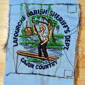 May include: Embroidered patch with a light blue background. The patch features a man standing on a boat, fishing. Text includes "LAFOURCHE PARISH SHERIFF'S DEPT" and "CAJUN COUNTRY" in a circular design. The image includes a church and trees.